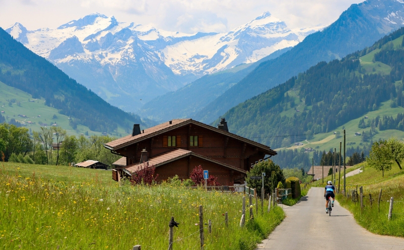 Cyclist riding alongside an alpine chalet with glacier mountains in the background