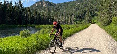 A solo gravel cyclist riding by a lake with tree-covered hills in background