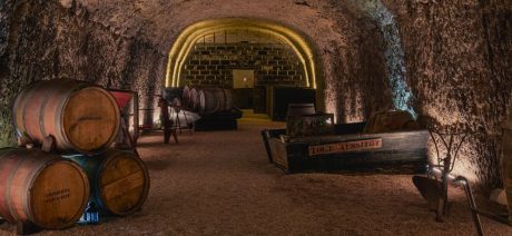 A dimly-lit troglodyte wine cellar with barrels and dirt floor.